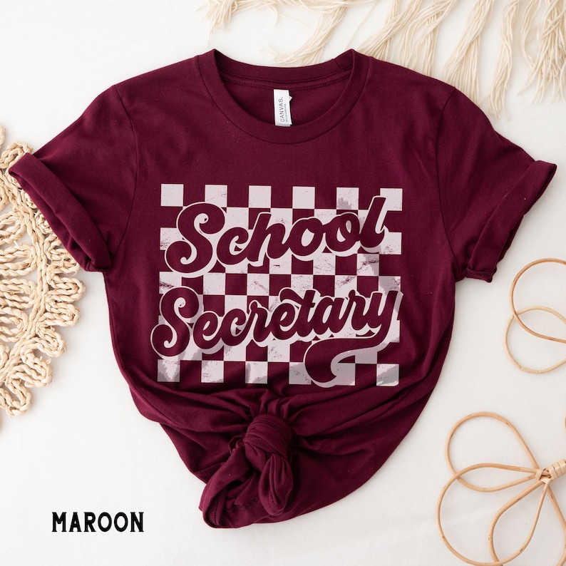 Peut inclure: T-shirt marron avec un fond &agrave; carreaux blancs et le texte "School Secretary" dans une police r&eacute;tro.