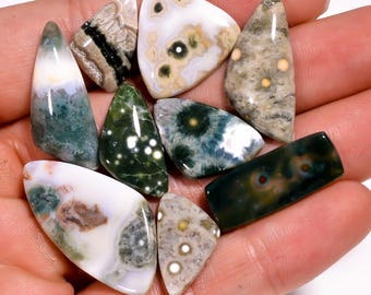 May include: A collection of polished gemstones in various shapes and colors, including green, white, brown, and beige. The gemstones are held in a hand, showcasing their unique patterns and textures. Ideal for jewelry making or collecting.