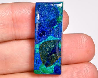 May include: A rectangular gemstone with a polished surface. The stone features a blend of deep blues, greens, and hints of gold. The colors are swirled and mottled, creating a unique pattern. The gemstone is held between fingers.