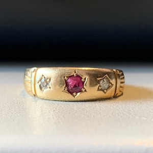 Puede incluir: Un anillo de banda de oro con un rubí rojo y dos pequeños diamantes engastados en forma de estrella.