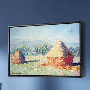 May include: A beige sofa with two pillows sits in front of a blue wall with a framed painting of haystacks in a field. A wooden coffee table with two drawers sits in front of the sofa.