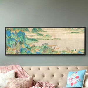 May include: A framed landscape painting depicting a serene lake scene with mountains and trees in the background. The painting is in a black frame and is hanging on a light blue wall above a beige sofa.