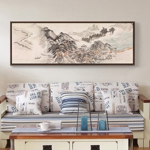 May include: A white couch with blue and white striped cushions and pillows with a black and white newspaper print. The couch is in front of a white wall with a framed black and white landscape painting above it. A white coffee table with two drawers is in front of the couch.