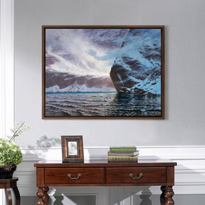 May include: A framed landscape painting depicts a dramatic coastal scene. The artwork features a large rock formation on the right, with a cloudy sky and ocean waves. The frame is dark brown, and the painting is mounted on a gray wall.