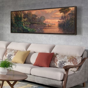 Puede incluir: Una pintura de paisaje enmarcada que representa una escena de río con árboles y una puesta de sol. La pintura está en un marco marrón y está colgada en una pared sobre un sofá beige con almohadas.