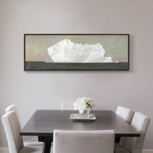 May include: A framed painting of a large iceberg floating in the ocean. The iceberg is white and the ocean is a dark blue. The painting is in a brown frame and is hanging on a white wall.