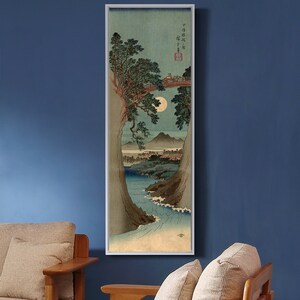 May include: A Japanese woodblock print depicting a moonlit scene with a bridge over a river. The print features a large tree with branches reaching across the river, and a mountain in the distance. The print is framed in a white frame.