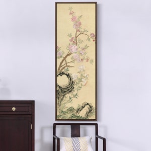 May include: A vertical painting of a delicate, stylized tree branch with pink and white blossoms on a pale yellow background. The tree trunk is a dark gray and the branches are thin and elegant. The painting is framed in a dark wood frame.