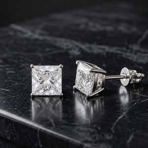 Puede incluir: Un par de pendientes de botón con talla princesa, con piedras cuadradas facetadas transparentes en un engaste de metal plateado de cuatro garras. Un pendiente se muestra de frente y el otro de lado, dejando ver el poste y el cierre.