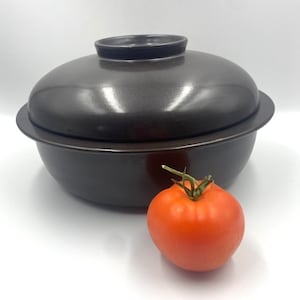 May include: A dark brown ceramic casserole dish with a lid, next to a ripe red tomato. The dish has a round shape with a lid that fits snugly on top. The tomato has a green stem.