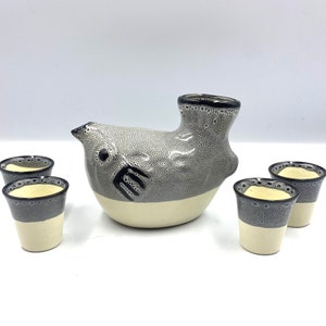 May include: A set of five ceramic sake cups and a ceramic sake bottle shaped like a bird. The bottle is gray and white with black accents. The cups are white with black rims.