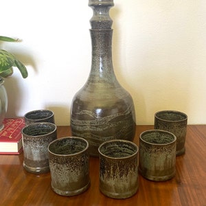 Puede incluir: Un juego de seis vasos de beber de cerámica marrón y verde con una jarra de cerámica marrón y verde a juego con una boca ancha y un cuello alto y estrecho.