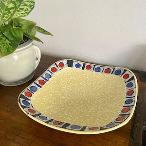 Może przedstawiać: Kwadratowa ceramiczna tacka do serwowania z żółtą, kropkowaną glazurą i czarnym, niebieskim i czerwonym geometrycznym wzorem wokół krawędzi.