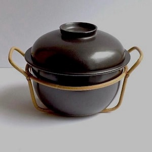May include: A dark brown ceramic cooking pot with a matching lid. The pot sits in a light brown rattan frame with handles. The pot is round with a slightly flared rim.
