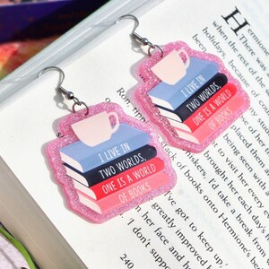 Può includere: Orecchini con glitter rosa con una pila di libri e un ciondolo a forma di tazza da caffè. I libri hanno il testo "Vivo in due mondi, uno è un mondo di libri".