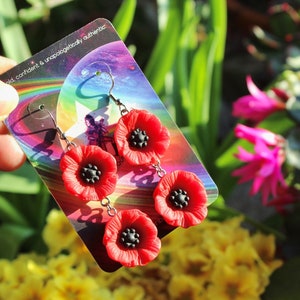 May include: A pair of red poppy flower dangle earrings with black centers. The earrings are on a card with the text "bold, confident & unapologetically authentic".