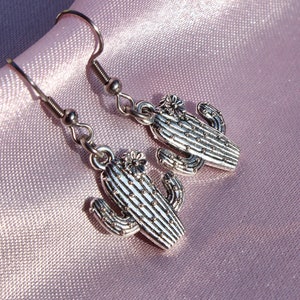 Op de afbeelding: Een paar zilveren cactus oorbellen met een bloemdetail. De oorbellen liggen op een roze satijnen achtergrond.