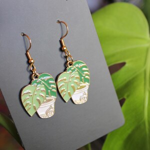 Op de afbeelding: Een paar goudkleurige oorhangers met een ontwerp van een potplant. Elke oorbel heeft een witte pot met een groene plant en goudkleurige accenten. De oorbellen worden gepresenteerd op een donkergrijze kaart.