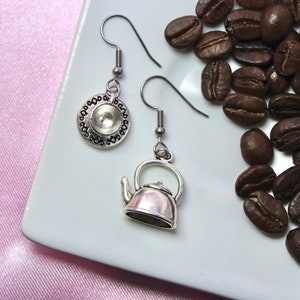Può includere: Orecchini d'argento con un ciondolo a forma di teiera e tazza da tè. Gli orecchini sono su un piatto bianco con chicchi di caffè.