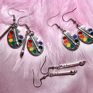 Op de afbeelding: Een paar zilveren oorbellen met verfpalette-charms en een paar zilveren oorbellen met penseel-charms. De oorbellen zijn op een roze pluche achtergrond.