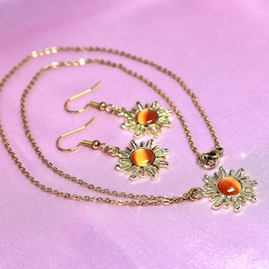 May include: A gold necklace and earrings set with a sun pendant and matching earrings. The sun features a red and orange gem in the center.