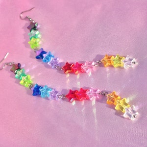 May include: Rainbow star-shaped dangle earrings with a silver chain. The earrings are made of iridescent plastic and have a rainbow colour scheme.