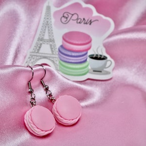 Könnte beinhalten: Rosa Macaron-förmige Ohrringe mit silbernen Haken. Die Ohrringe befinden sich auf einem rosa Satin-Hintergrund mit einem Aufkleber des Eiffelturms und dem Wort "Paris".