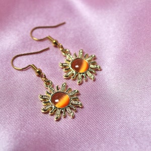 May include: A pair of gold earrings with a sun design and orange gemstone accents.