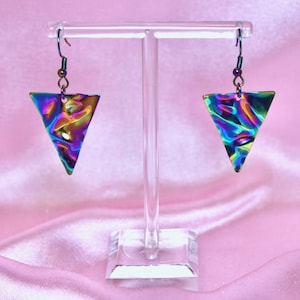 May include: A pair of iridescent triangle earrings with a textured surface. The earrings are hanging from a clear display stand.