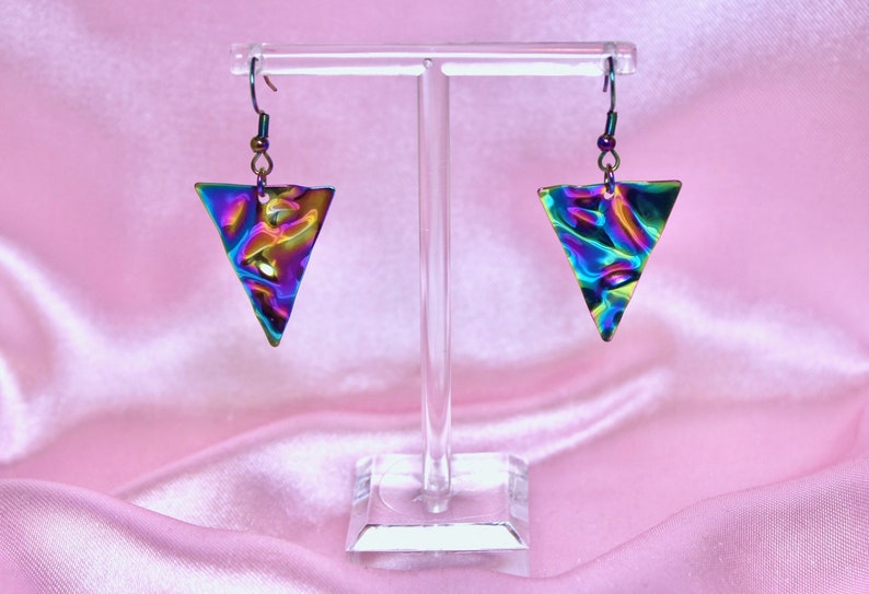 May include: A pair of iridescent triangle earrings with a textured surface. The earrings are hanging from a clear display stand.