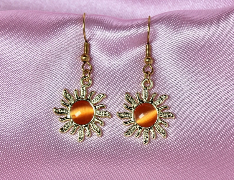 May include: A pair of gold sun earrings with orange gemstone centers. The earrings have a delicate, detailed design.