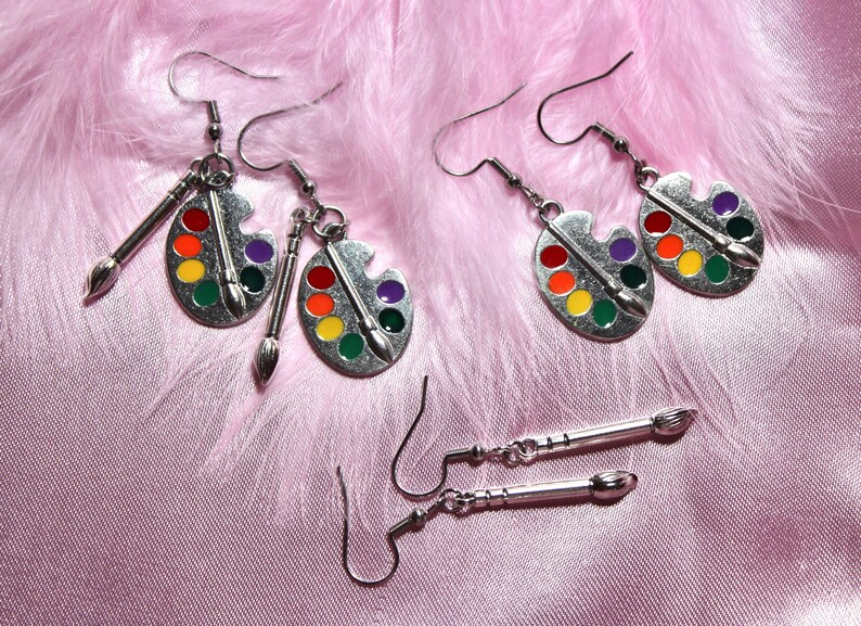 Op de afbeelding: Een paar zilveren oorbellen met verfpalette-charms en een paar zilveren oorbellen met penseel-charms. De oorbellen zijn op een roze pluche achtergrond.