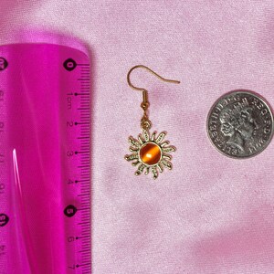 May include: Gold sun-shaped earring with an orange gemstone in the center. The earring is hanging from a gold hook.