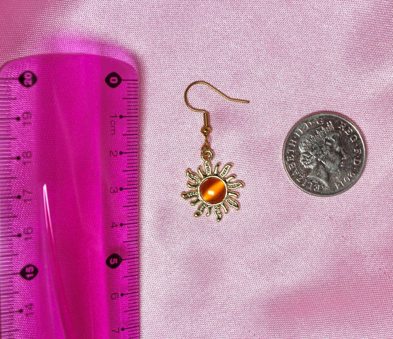 May include: Gold sun-shaped earring with an orange gemstone in the center. The earring is hanging from a gold hook.