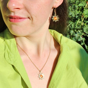 May include: A gold necklace with a sun pendant featuring an orange stone. The necklace is worn by a person with a green shirt and matching gold earrings with sun pendants.