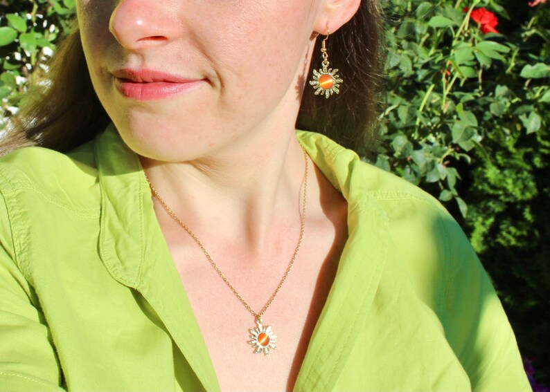 May include: A gold necklace with a sun pendant featuring an orange stone. The necklace is worn by a person with a green shirt and matching gold earrings with sun pendants.