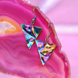 May include: A pair of iridescent triangle earrings with silver hooks. The earrings are on a pink and white agate background.