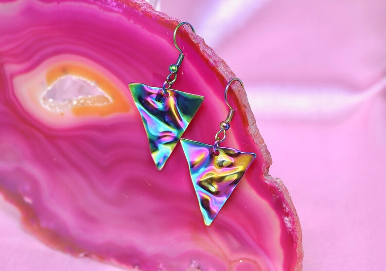 May include: A pair of iridescent triangle earrings with silver hooks. The earrings are on a pink and white agate background.