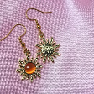 May include: A pair of gold sun earrings with orange glass stones. The earrings have a delicate, intricate design.