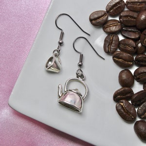 Könnte beinhalten: Ein Paar silberne Ohrringe mit einem Teekannen- und einem Teetassen-Anhänger. Die Ohrringe liegen auf einer weißen Oberfläche mit Kaffebohnen, die um sie herum verstreut sind.