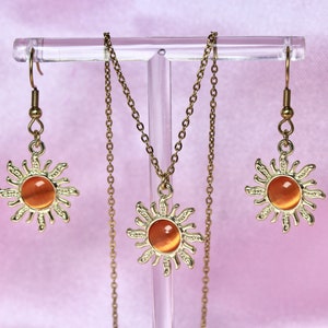 May include: A gold necklace and earrings set featuring a sun design with an orange gemstone in the center. The necklace and earrings are displayed on a clear stand.
