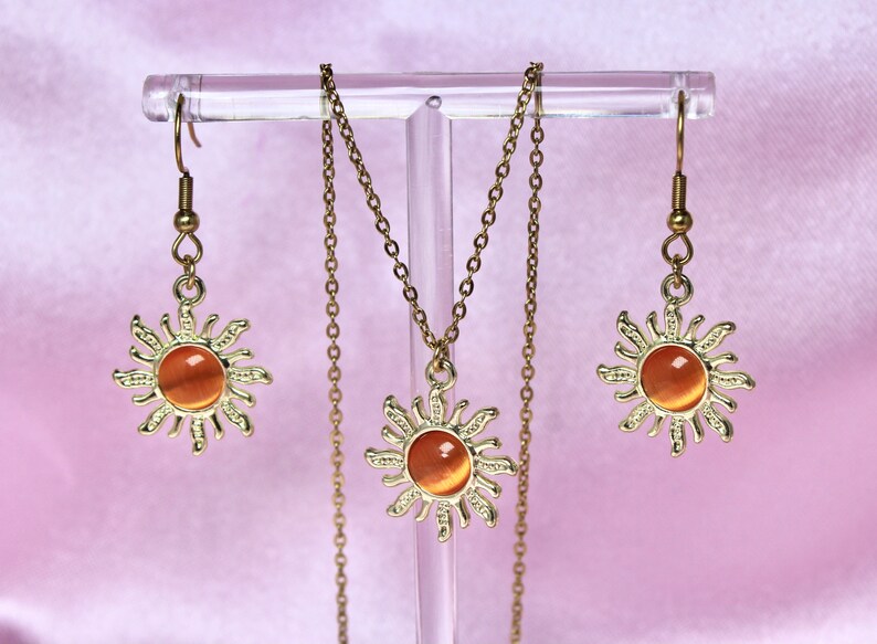 May include: A gold necklace and earrings set featuring a sun design with an orange gemstone in the center. The necklace and earrings are displayed on a clear stand.