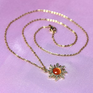 May include: A gold chain necklace with a sun pendant featuring an orange gemstone. The necklace is on a pink fabric background.