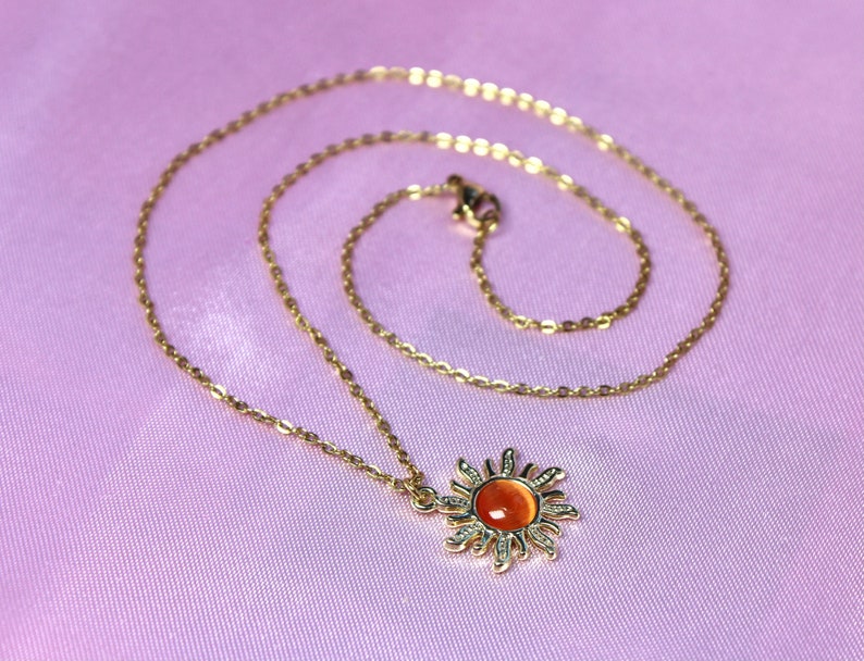May include: A gold chain necklace with a sun pendant featuring an orange gemstone. The necklace is on a pink fabric background.