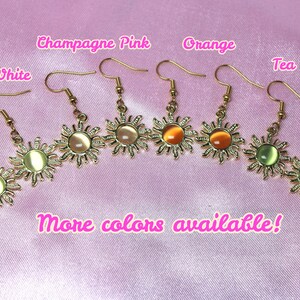 May include: Gold sun earrings with different colored glass stones. The colors are ivory white, champagne pink, orange, tea green. The text "More colors available!" is written below the earrings.