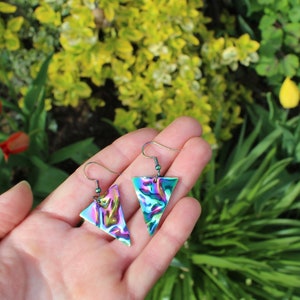 May include: A pair of iridescent triangle earrings with a silver hook. The earrings have a marbled pattern of green, blue, and purple.