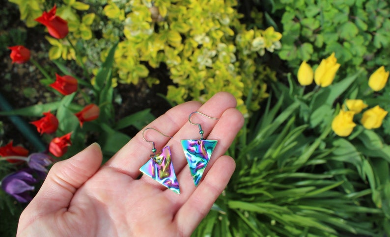May include: A pair of iridescent triangle earrings with a silver hook. The earrings have a marbled pattern of green, blue, and purple.