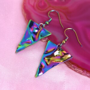 May include: A pair of iridescent triangle earrings with a hammered metal finish. The earrings are on a pink background.
