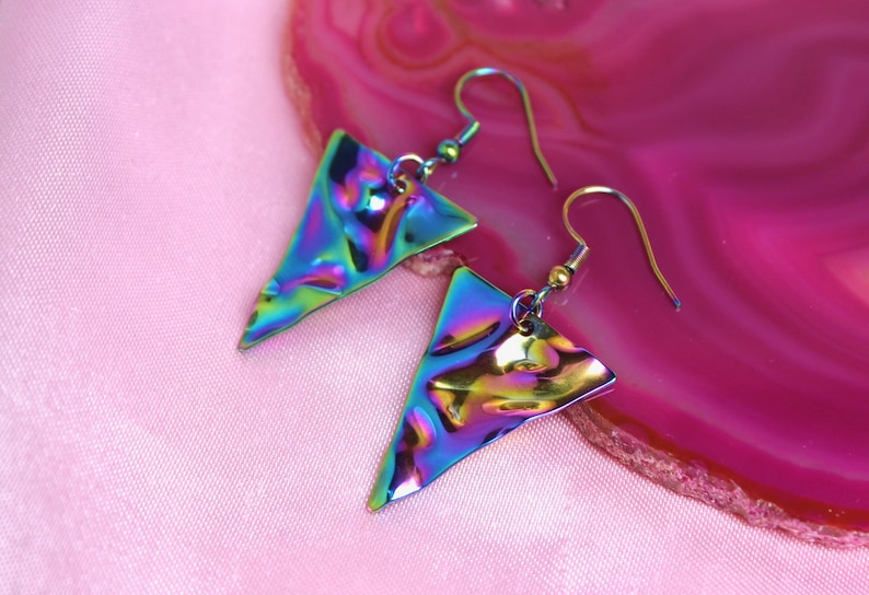 May include: A pair of iridescent triangle earrings with a hammered metal finish. The earrings are on a pink background.