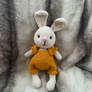 May include: A handmade crochet bunny doll with a white body, long ears, and a pink nose. The bunny wears a mustard yellow jumpsuit. The doll is sitting on a soft, gray, textured surface.